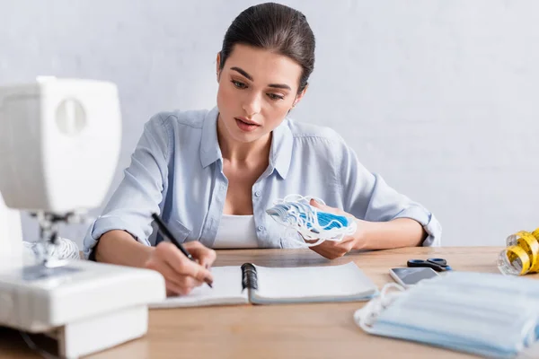 Seamstress writing on notebook near medical masks and blurred sewing machine — Stock Photo