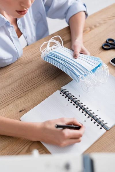Cropped view of seamstress holding medical masks and writing on notebook — Stock Photo