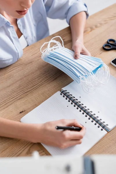 Ausgeschnittene Ansicht einer Näherin, die medizinische Masken hält und auf ein Notizbuch schreibt — Stockfoto