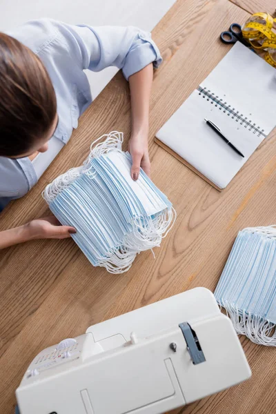 Vista aérea de costurera sosteniendo máscaras médicas cerca de portátil y máquina de coser - foto de stock