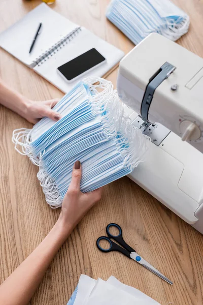 Ausgeschnittene Ansicht medizinischer Masken in den Händen einer Näherin, die in der Nähe von Nähmaschine und Smartphone arbeitet — Stockfoto