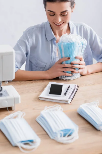 Costurera sonriente sosteniendo máscaras médicas cerca del teléfono celular y la máquina de coser - foto de stock