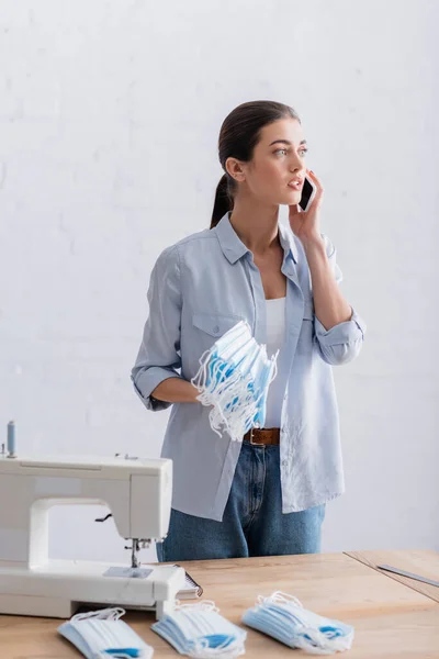 Seamstress with medical masks talking on smartphone near sewing machine — Stock Photo