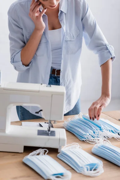 Cropped view of young seamstress talking on smartphone near sewing machine and medical masks on blurred foreground — Stock Photo