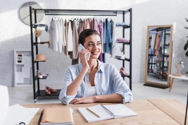 Smiling designer talking on smartphone near notebooks on table — Stock Photo