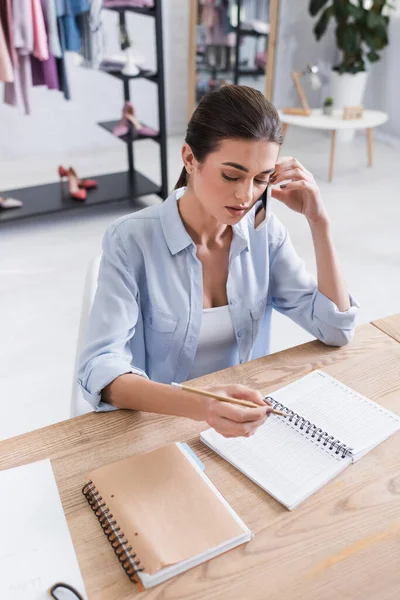 Jeune couturière écrivant sur carnet et parlant sur téléphone portable en atelier — Photo de stock