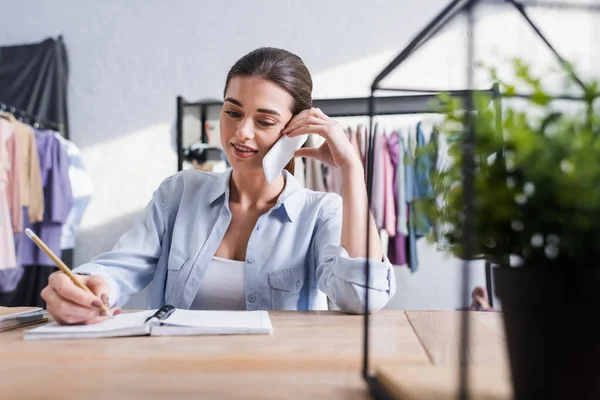 Seamstress falando no smartphone e escrevendo no notebook perto da planta turva — Fotografia de Stock