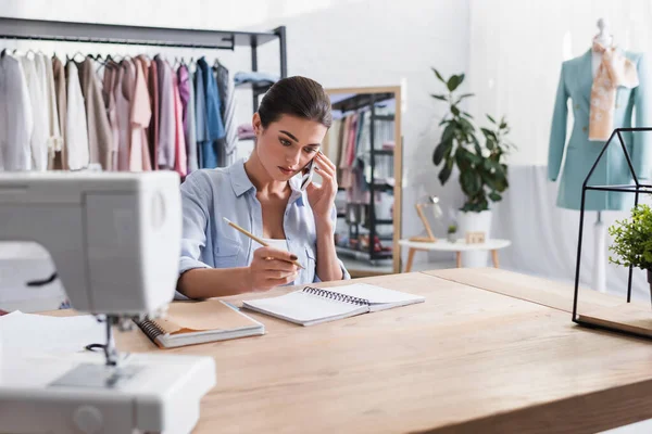 Diseñador hablando en el teléfono inteligente cerca de la máquina de coser borrosa y portátiles - foto de stock