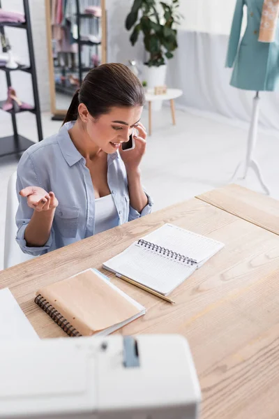 Cheerful seamstress talking on mobile phone near notebooks in atelier — Stock Photo