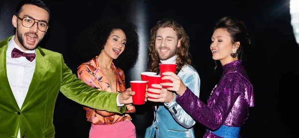 Amigos multirraciais positivos em roupas coloridas bebendo de copos de plástico em boate no fundo preto, banner — Fotografia de Stock