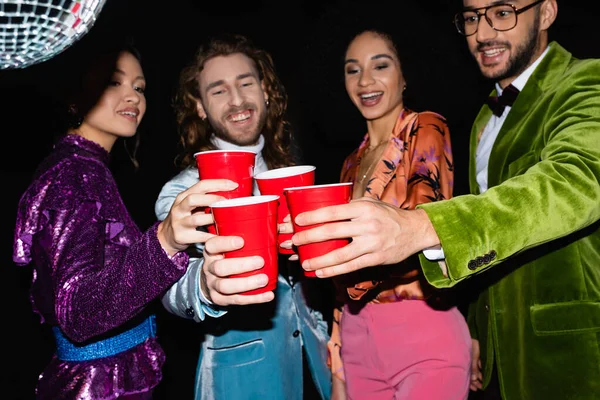 Улыбающиеся многорасовые друзья в разноцветной одежде тост с пластиковыми чашками в ночном клубе на черном фоне — стоковое фото