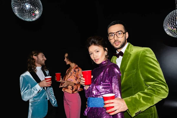 Amigos multirraciais positivos em roupas coloridas de pé com copos de plástico nas mãos sobre fundo preto — Fotografia de Stock