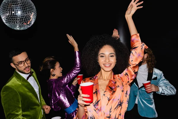 Positive afrikanisch-amerikanische Frau tanzt auf Party mit multirassischen Freunden in bunten Kleidern auf schwarzem Hintergrund — Stockfoto