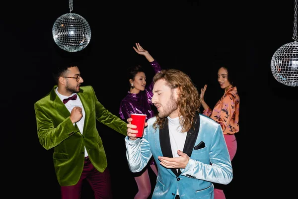 Giocoso giovane adulto in possesso di tazza di plastica e smorfia sulla festa con amici multirazziali che ballano su sfondo nero — Foto stock