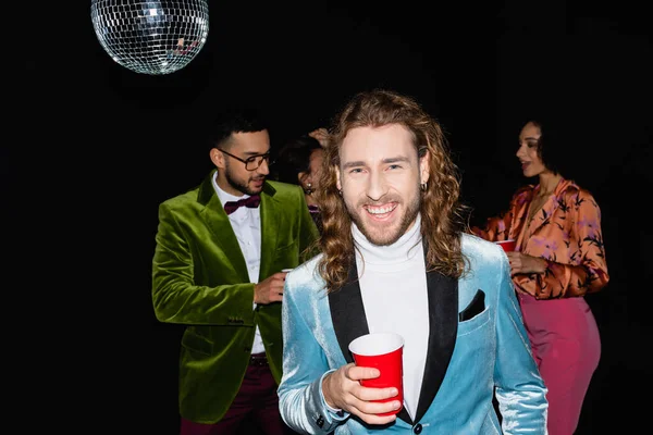 Sonriente joven adulto hombre sosteniendo taza de plástico cerca borrosa multirracial amigos de pie sobre fondo negro - foto de stock