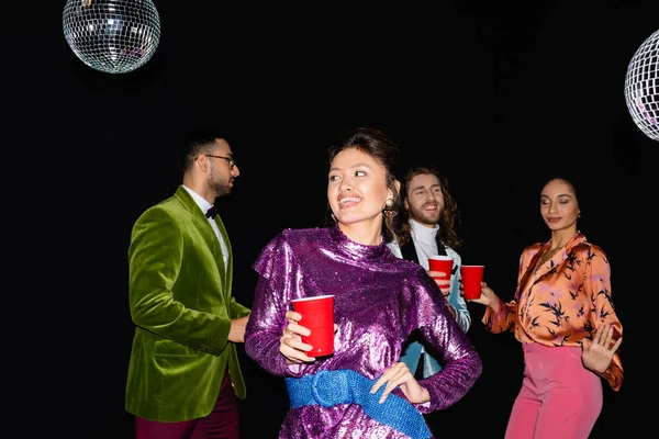 Verspielte junge erwachsene asiatische Frau, die in der Hand auf Hüfthöhe in der Nähe von interrassischen Freunden steht, die auf schwarzem Hintergrund tanzen — Stockfoto