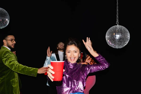 Asiático mujer con plástico taza bailando cerca borrosa multiracial amigos en colorido ropa en negro fondo - foto de stock