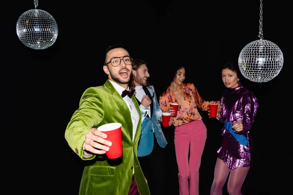 Homme arabe positif dans des lunettes de danse avec des amis multiraciaux en vêtements colorés dans une boîte de nuit sur fond noir — Photo de stock