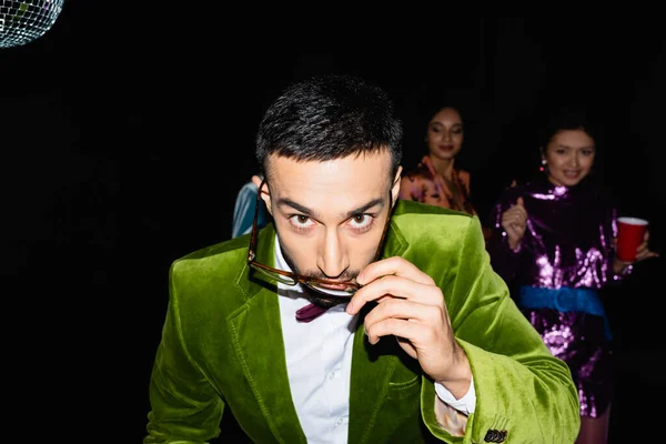 Young adult arab man adjusting glasses while having party with multiracial friends on black background — Stock Photo