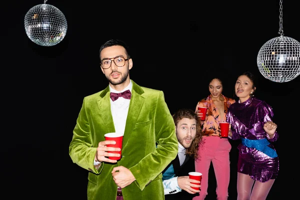 Young adult arab man standing near funny multiracial friends in colorful clothes at party on black background — Stock Photo