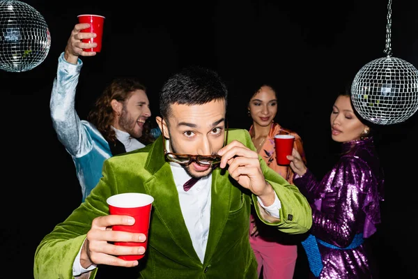 Young adult arab man adjusting glasses near funny multiracial friends in colorful clothes at party on black background — Stock Photo