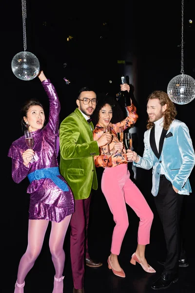 Verspielte multirassische Freunde in bunten Kleidern mit Champagnergläsern in den Händen, die auf schwarzem Hintergrund tanzen — Stockfoto
