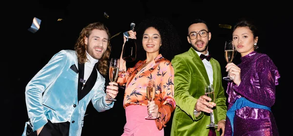 Allegri amici multirazziali che ballano con bicchieri di champagne su sfondo nero, striscione — Foto stock