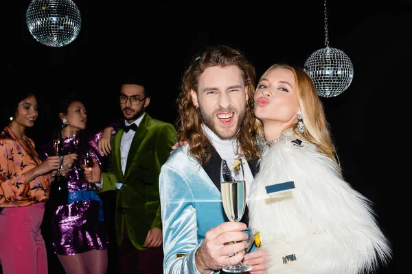 Pareja positiva haciendo muecas en la fiesta con amigos multirraciales en el fondo negro - foto de stock