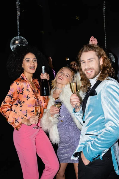 Positive multirassische Freunde in bunten Kleidern tanzen mit Champagner in Nachtclub auf schwarzem Hintergrund — Stockfoto