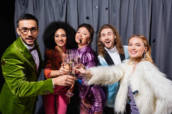 Amis interracial dans des vêtements élégants cliquetis avec des verres de champagne près du rideau gris sur fond noir — Photo de stock