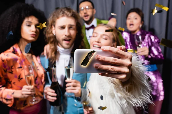 Amigos inter-raciais positivos em roupas brilhantes tomando selfie no smartphone perto de cortina cinza no fundo preto — Fotografia de Stock