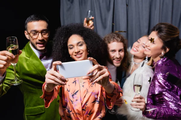 Amigos inter-raciais em roupas brilhantes tomando selfie no smartphone perto de cortina cinza no fundo preto — Fotografia de Stock