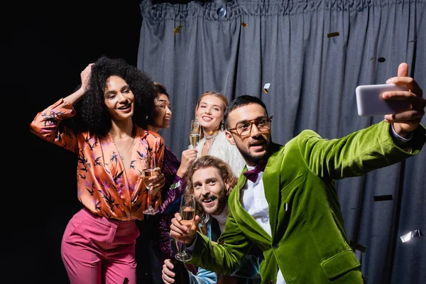Stylish interracial friends in bright clothes taking selfie on smartphone near grey curtain on black background — Stock Photo