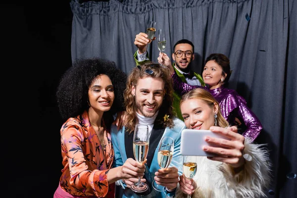 Sonriente interracial amigos en brillante ropa tomando selfie en smartphone cerca gris cortina en negro fondo - foto de stock