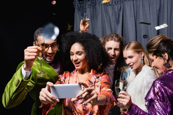 Positive interracial friends in bright clothes taking selfie on smartphone near grey curtain on black background — Stock Photo