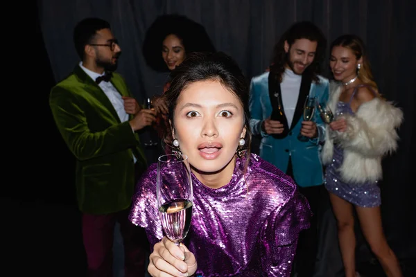 Asiatische junge erwachsene Frau blickt in die Kamera und hält ein Glas Champagner neben verschwommenen interrassischen Freunden auf grauem Hintergrund — Stockfoto