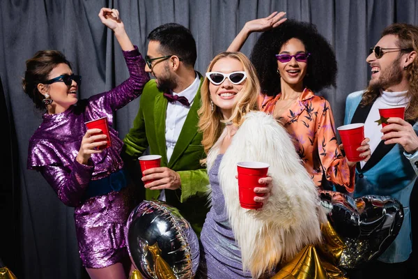 Amigos interracial divertidos en ropa colorida con copas de plástico en las manos bailando sobre fondo gris — Stock Photo