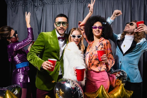 Positive interracial friends in colorful clothes and sunglasses dancing near grey curtain on black background — Stock Photo