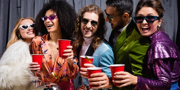 Amigos interraciales juguetones en gafas de sol celebrando con copas de plástico sobre fondo gris, pancarta - foto de stock