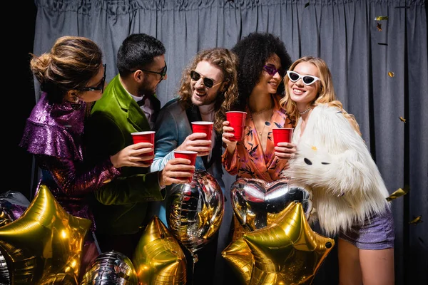 Felices amigos interracial en gafas de sol celebrando con copas de plástico cerca de cortina gris sobre fondo negro — Stock Photo