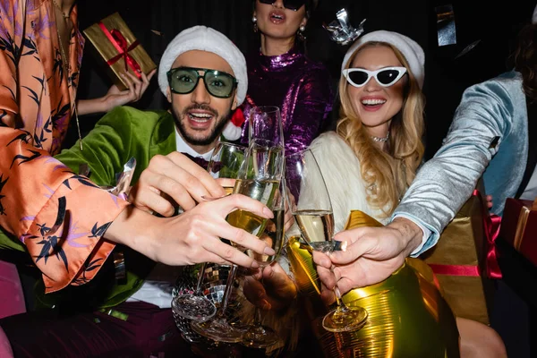 Lächelnde interrassische Freunde in Weihnachtsmützen und Sonnenbrillen, die mit Champagnergläsern auf schwarzem Hintergrund klirren — Stockfoto