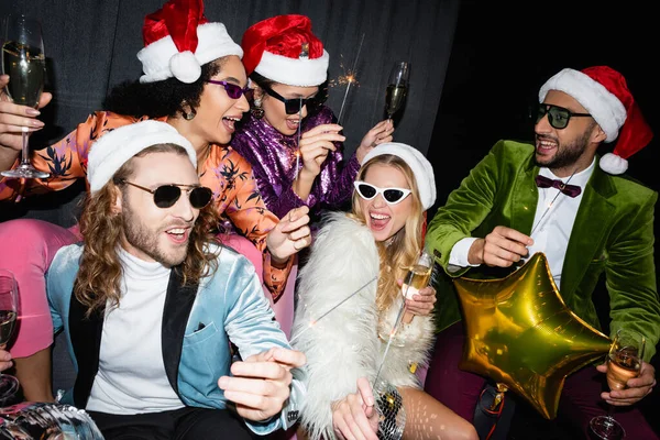 Межрасовые друзья в шляпах Санты пьют шампанское и празднуют Новый год возле серого занавеса на черном фоне — стоковое фото