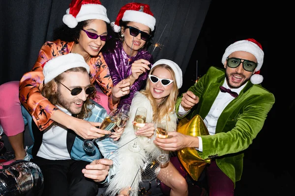 Amigos inter-raciais brincalhões em chapéus de santa celebrando o ano novo perto cortina cinza no fundo preto — Fotografia de Stock