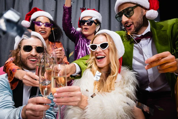 Улыбающиеся межрасовые друзья в шляпах Санты, празднующие Новый год возле серого занавеса на черном фоне — стоковое фото