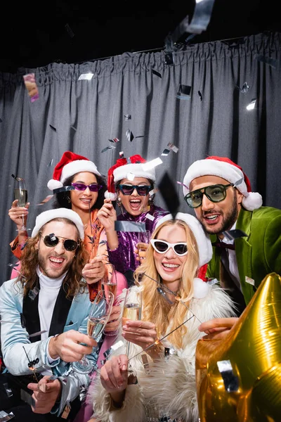 Stylish interracial friends in santa hats and sunglasses having party near grey curtain on black background — Stock Photo