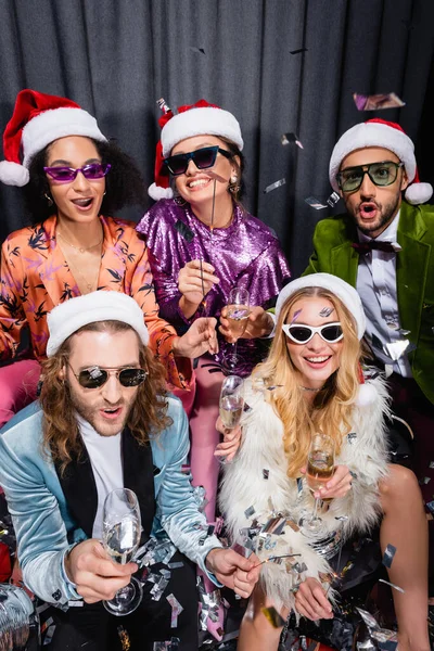 Jóvenes adultos interracial amigos en santa sombreros y gafas de sol teniendo fiesta en gris fondo — Stock Photo