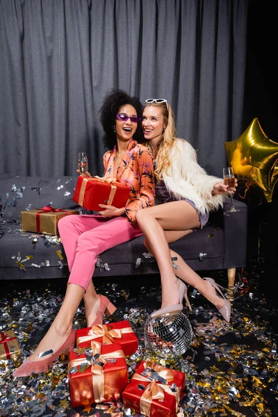 Happy young adult interracial women sitting with champagne on grey couch near curtain on black background — Stock Photo