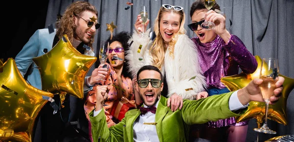 Fröhliche interrassische Freunde in Sonnenbrille und farbenfroher Kleidung, Champagner trinkend neben grauem Vorhang auf schwarzem Hintergrund, Banner — Stockfoto