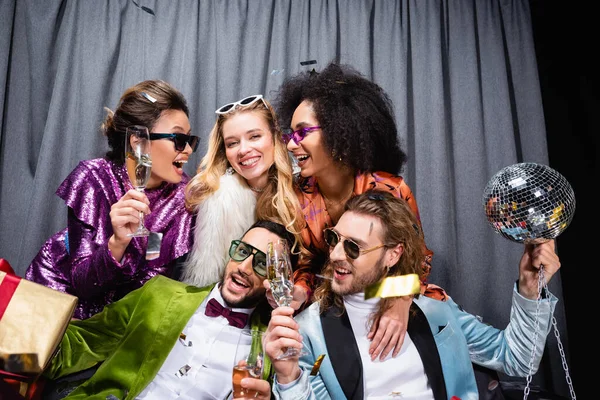 Amigos inter-raciais brincalhões em roupas coloridas bebendo champanhe perto cortina cinza no fundo preto — Fotografia de Stock