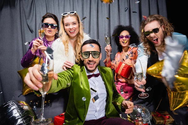 Souriant amis interracial en vêtements colorés boire du champagne près du rideau gris sur fond noir — Photo de stock